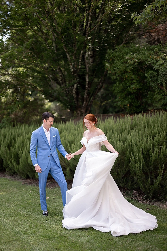Couple portrait of bride and groom holding hands, her off-the-shoulder gown with long train, his blue suit, on a garden lawn with hedges