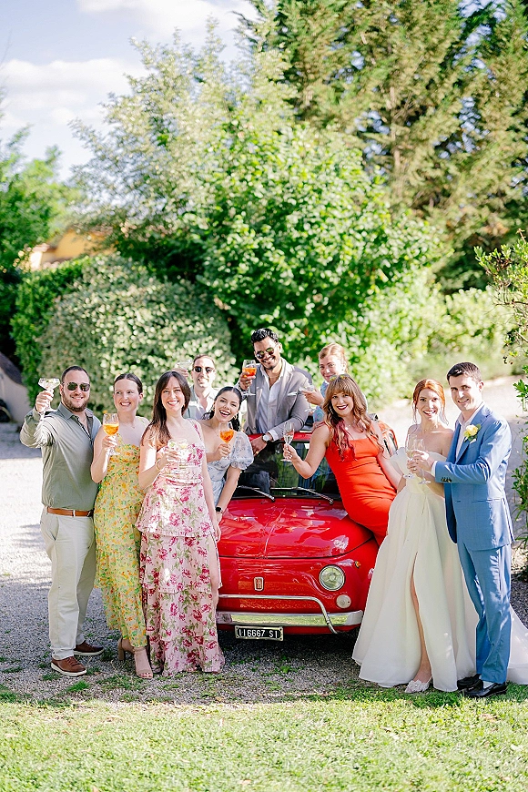 Wedding party portrait with drinks as bride and groom toast with friends in sunglasses beside a red vintage car on a sunny driveway