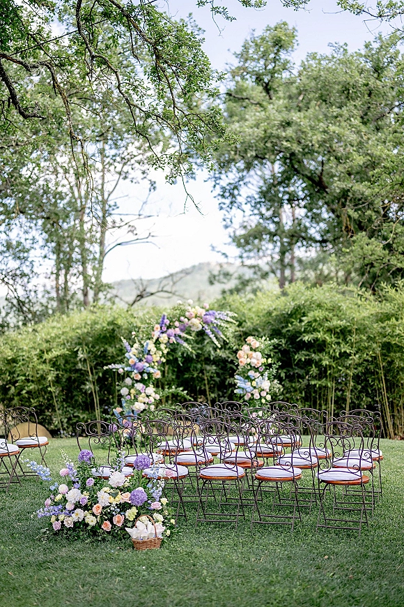 Outdoor ceremony setup with a garden wedding ceremony floral arch, pastel aisle flowers, wrought iron chairs, on a green lawn with trees and hills