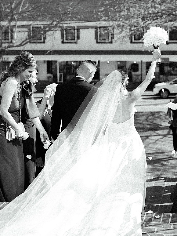 Wedding recessional as bride and groom exit holding hands, bride lifting bouquet overhead with long veil train on brick walkway street