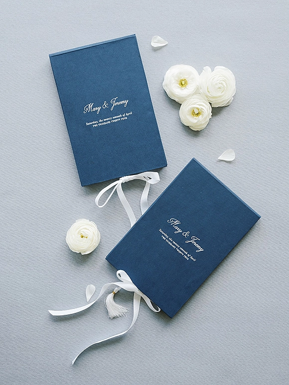 Wedding invitation suite with navy wedding invitations tied in white ribbon with tassel and white flowers on a light gray fabric backdrop