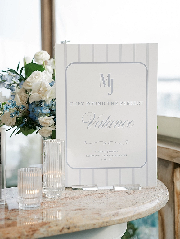 Wedding welcome sign in an acrylic holder on a round stone table with ribbed glass candles, white roses and blue hydrangea by a sunlit window view