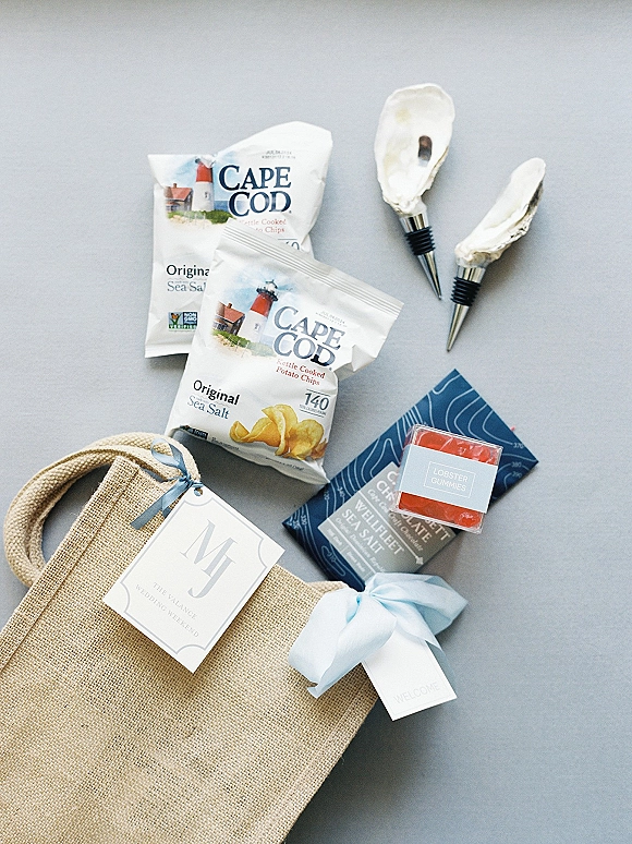 Wedding welcome bag flat lay featuring a burlap tote with blue ribbon, gift tags, snacks, chocolate, candy, and oyster shell stoppers on gray surface