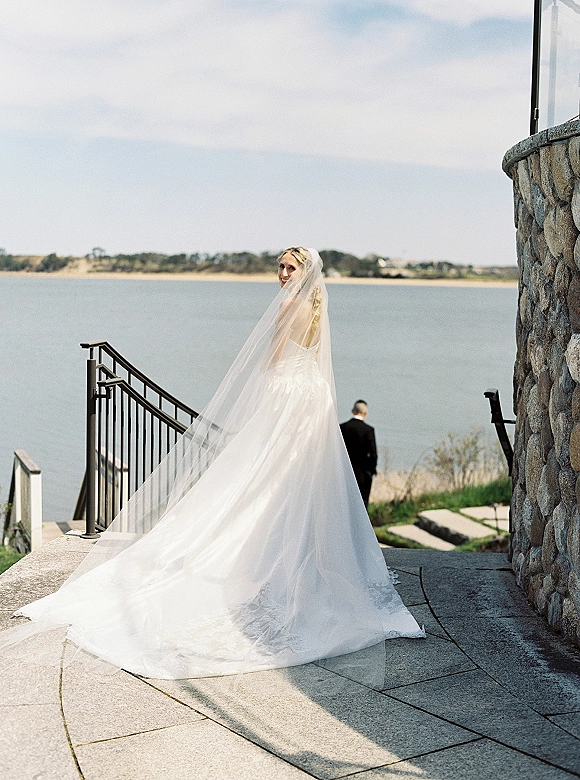 Bridal portrait of a bride in a strapless ball gown with cathedral veil blowing in the wind on a stone terrace by the waterfront
