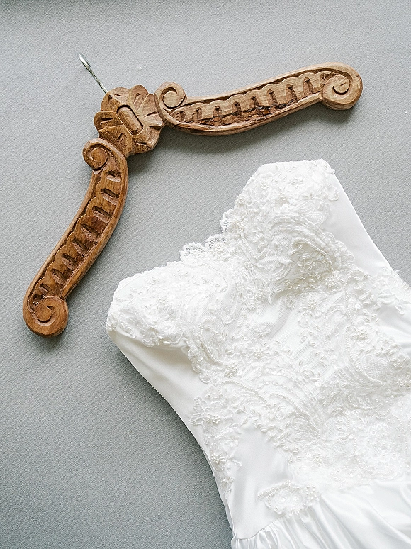 Wedding dress close-up of a strapless wedding dress bodice with lace applique and beadwork on a carved wooden hanger against gray fabric backdrop