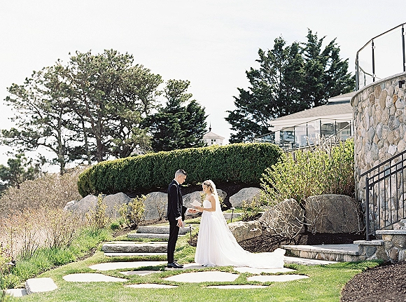 Wedding first look as bride in veil and wedding dress meets groom in black tuxedo on stone steps beside a green garden lawn