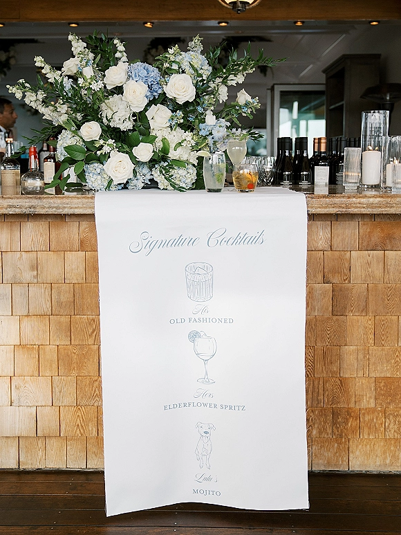Signature cocktail sign with wedding signature cocktails, white roses and blue hydrangeas, candles, and glasses on a rustic indoor bar counter