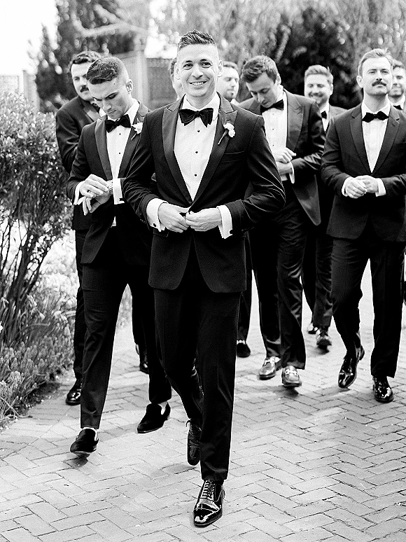 Groom with groomsmen in black tuxedos and bow ties walking on a brick paver walkway with greenery and hedges behind