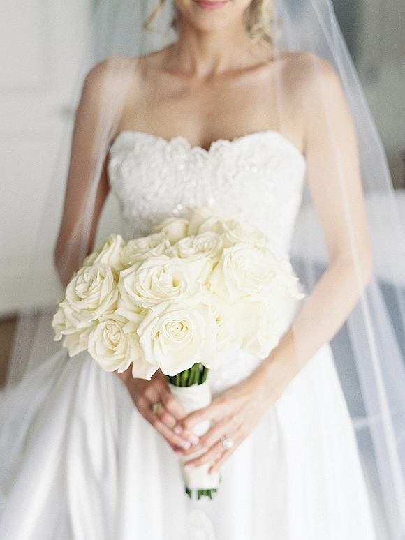 Bridal bouquet of white roses held by a bride in a strapless lace dress, veil over shoulders, engagement ring visible indoors