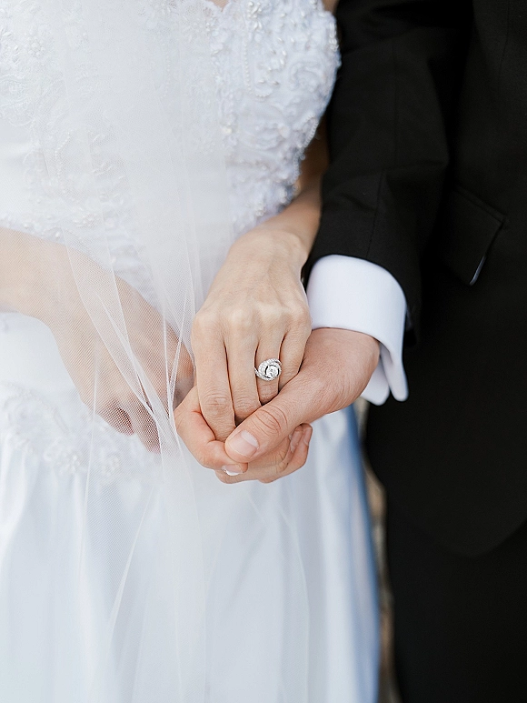 Wedding ring close-up of a diamond halo engagement ring on the bride’s hand, with lace dress and groom tuxedo cuff softly blurred behind