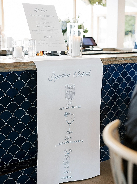 Signature cocktails sign on an acrylic stand with illustrated drinks and pillar candles in glass hurricanes on a blue scallop tile bar