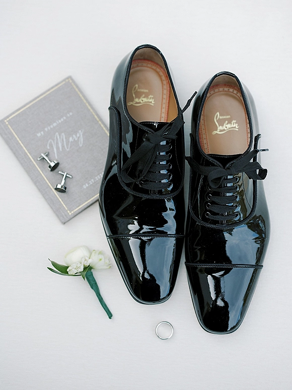 Groom shoes in black patent dress shoes on white fabric with wedding ring, cufflinks, boutonniere, and vow book flat lay