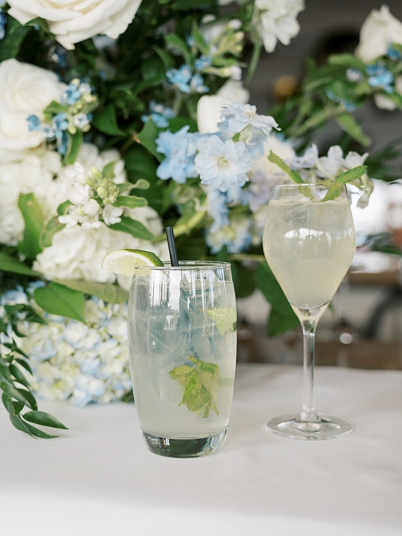 Wedding signature drinks in cocktail glasses with black straws, lime and mint garnishes beside blue and white flowers on a reception table