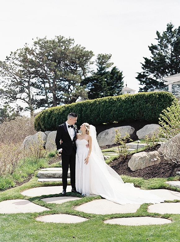 Couple portrait of bride and groom holding each other, her long veil trailing over a garden lawn beside a stepping stone path