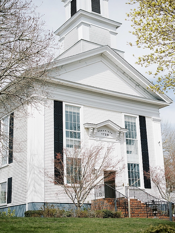 Church exterior with a white church facade, black shutters, and tall steeple above brick entrance steps with railing and trees beyond