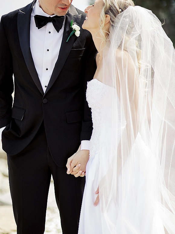 Couple portrait of bride and groom holding hands, she in strapless dress with veil, he in tuxedo with white boutonniere outdoors