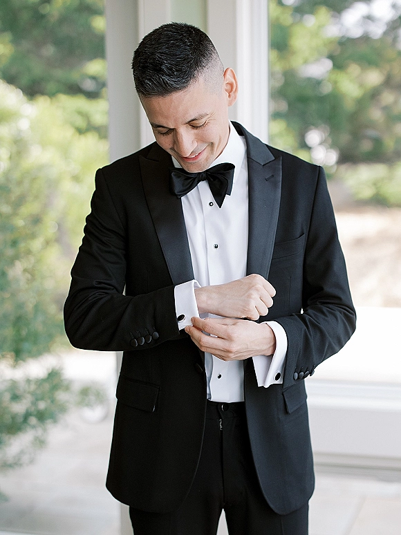Groom portrait in window light, wearing a black tuxedo with satin lapel and bow tie, adjusting cufflinks beside greenery and trees