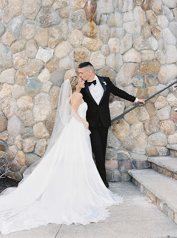 Couple portrait of groom kissing bride’s forehead as she stands in a strapless gown with long veil and lace train by stone steps