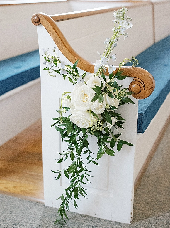 Ceremony pew flowers with white blooms and greenery spray attached to a church pew by a wood banister along the carpeted aisle