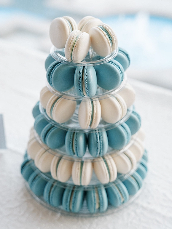Macaron tower with blue and white macarons stacked on a clear tiered stand atop a white tablecloth in soft blurred light background