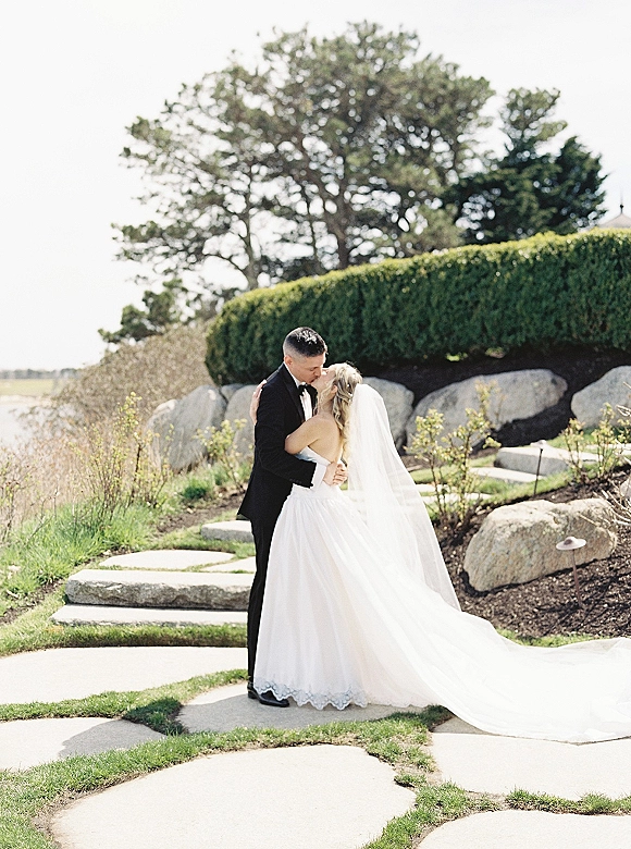 Wedding kiss portrait of bride and groom kissing on a garden path, her long veil and dress train flowing beside his black tuxedo