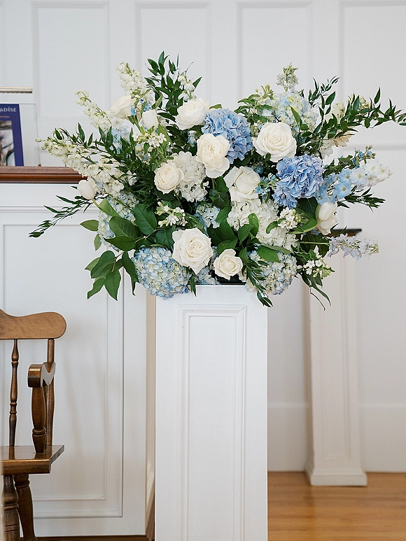 Wedding ceremony flowers on a white pedestal with white roses and blue hydrangeas, lush greenery, set against a white paneled wall