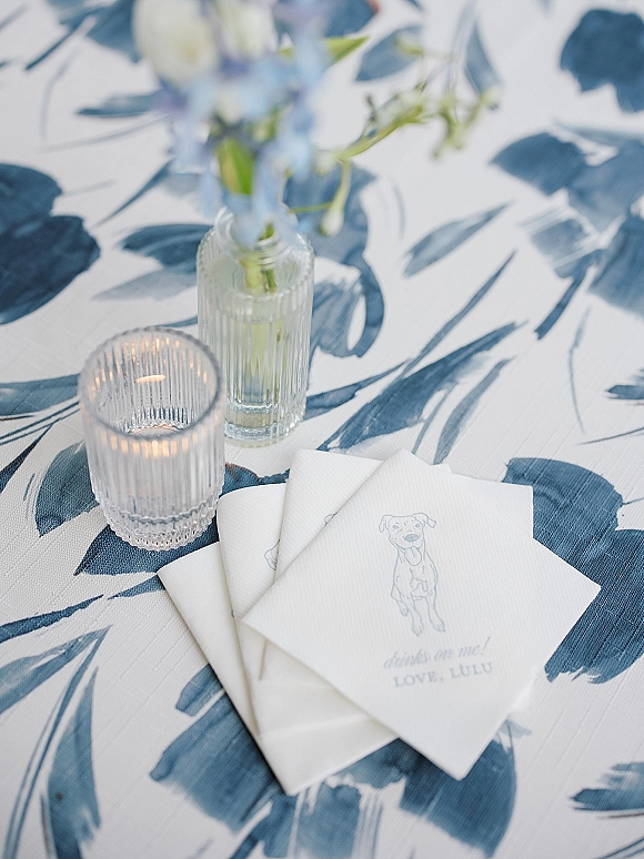 Wedding table detail with blue and white table linen, paper cocktail napkins, and a lit ribbed glass votive beside blue flowers
