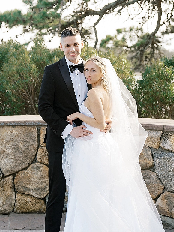 Couple portrait of bride and groom pose, her long veil draped over a strapless gown as he hugs her in front of a stone wall terrace