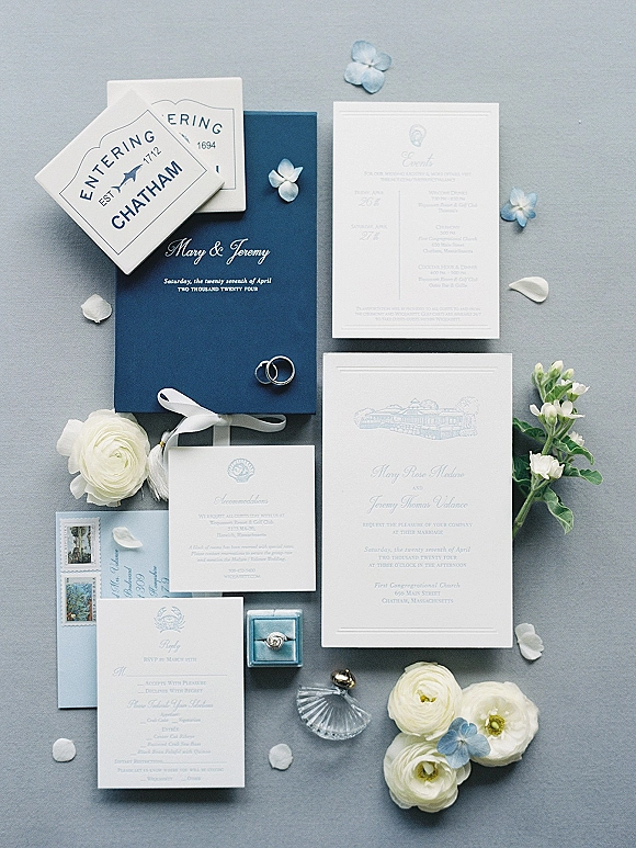 Invitation suite with blue wedding invitations, wax seal and vintage postage, rings in a box, and petals on a gray fabric backdrop