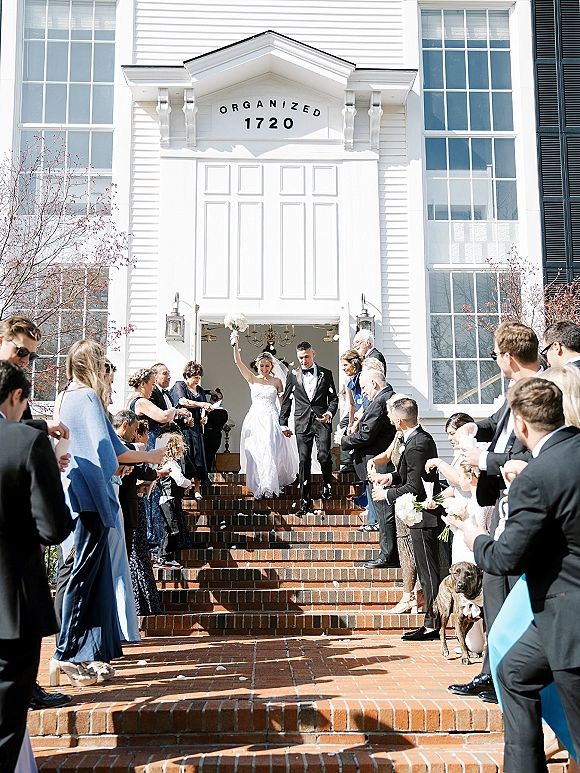 Wedding recessional with newlyweds walking down aisle as guests toss confetti, bride lifting bouquet on brick church steps outside a white facade