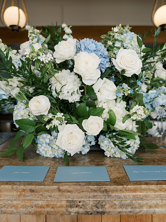 Wedding floral arrangement with white and blue centerpiece of roses, hydrangea, and greenery beside blue place cards on a stone countertop