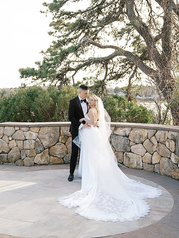 Couple portrait of bride and groom embrace, her lace train and cathedral veil flowing as he holds her waist on a stone terrace outdoors