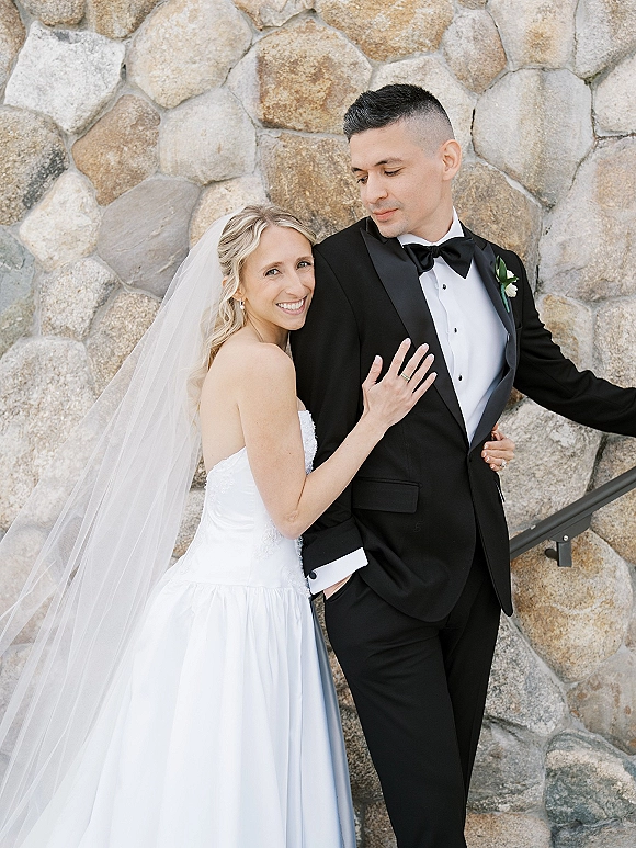 Couple portrait of bride and groom, bride hugging groom and holding his lapel as he looks at her beside a stone wall railing