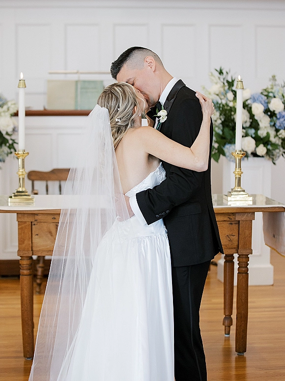 Wedding kiss portrait of bride and groom kissing at a church altar, her long veil and strapless dress beside candles and flowers