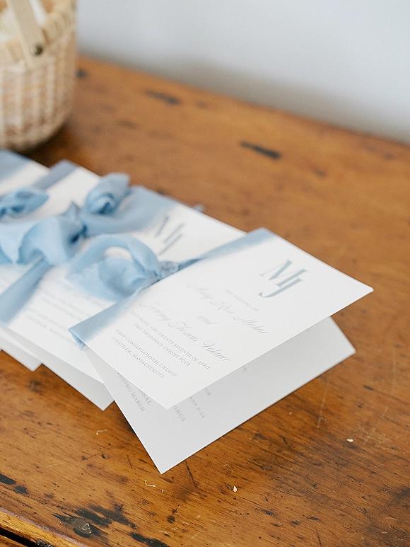 Invitation suite with blue silk ribbon, envelopes, and printed cards styled in a flat lay on a wood table with wicker basket backdrop