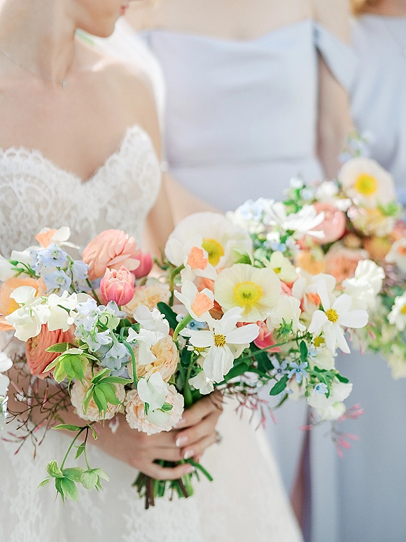 Bridal bouquet of white cosmos, peach roses, pink ranunculus, and blue delphinium held against a lace wedding dress in soft indoor light