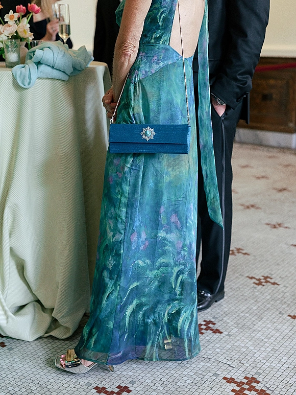 Wedding guest outfit in a teal printed dress holding a blue clutch purse beside a cocktail table with champagne and floral centerpiece indoors