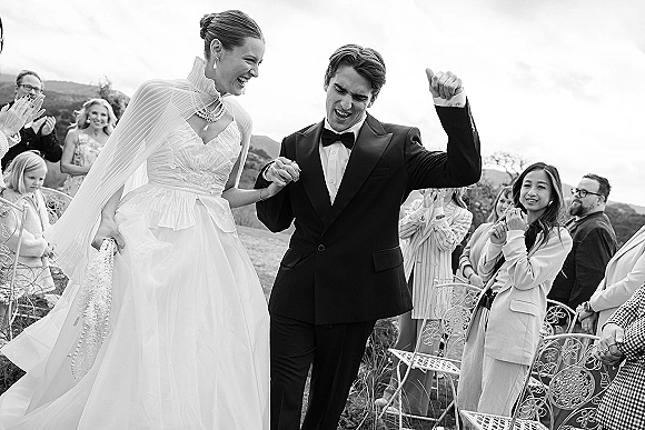 Recessional moment as bride and groom walk hand in hand down the aisle, bride in cape veil, groom fist pumping, guests applauding outdoors