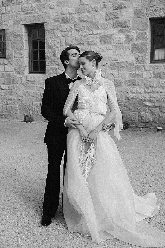 Wedding couple portrait in black and white wedding photo as groom kisses bride’s forehead, her pearl necklace and sheer cape by a stone wall
