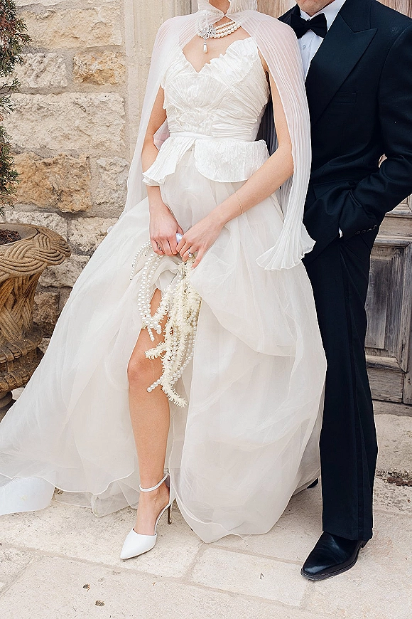 Bride and groom portrait in black tie attire, bride in strapless gown with pleated cape and pearl purse beside a stone doorway