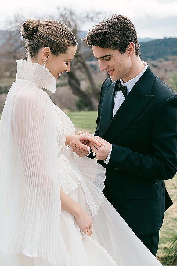 Wedding couple portrait of groom placing ring on bride’s hand, her pleated cape and ruffle collar visible against rolling hills outdoors