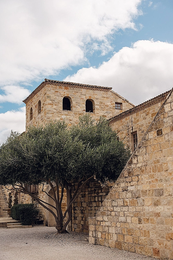 Stone wedding venue with rustic stone venue exterior, arched windows and clay tile roof, overlooking a gravel courtyard with an olive tree under blue sky