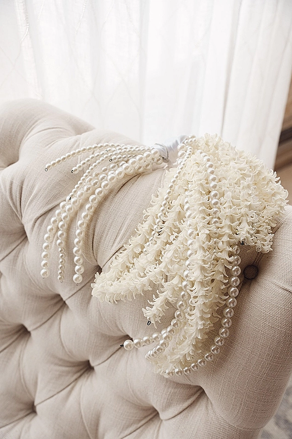 Bridal bouquet of all-white flowers with pearl strands draped over it, resting on a tufted chair beside sheer curtains