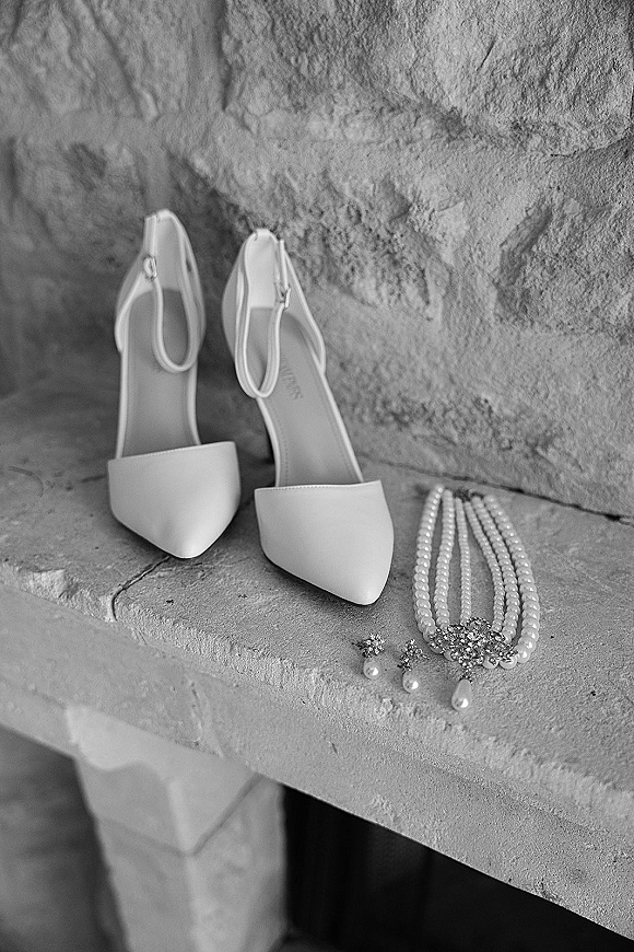 Bridal shoes and white wedding heels with ankle straps arranged beside pearl necklace and drop earrings on a stone ledge against textured wall