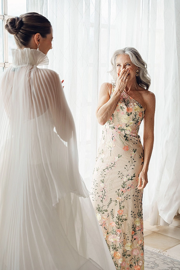 Bride reveal moment as mom turns in surprise, bride in veil and gown with pearl drop earrings by sheer curtains in window light indoors