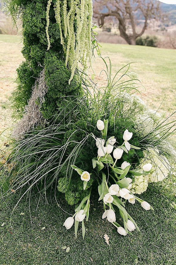 Wedding ceremony florals on a moss covered ceremony pillar with hanging amaranthus and white tulips, set on a grassy field with trees