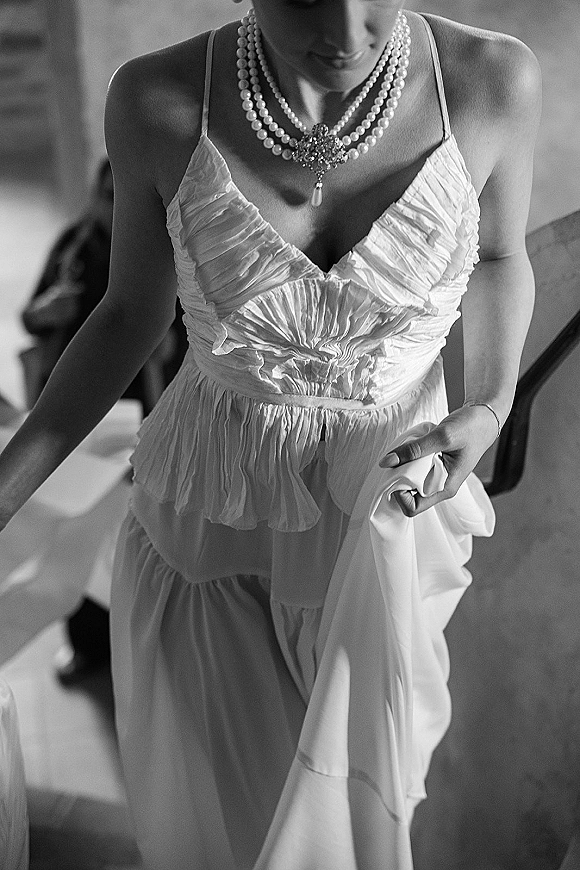 Bridal gown close-up with pearl wedding necklace and brooch pendant over ruched bodice and spaghetti straps in a softly blurred indoor room