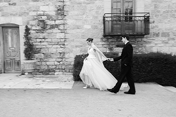 Couple portrait of bride in a strapless wedding dress with cape leading groom by the hand past a stone wall and wooden door courtyard
