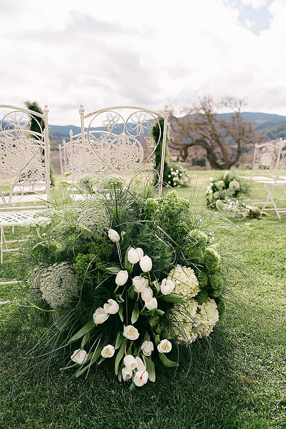 Ceremony aisle decor with outdoor ceremony chairs and low tulip, hydrangea, and greenery clusters on a lawn with distant hills