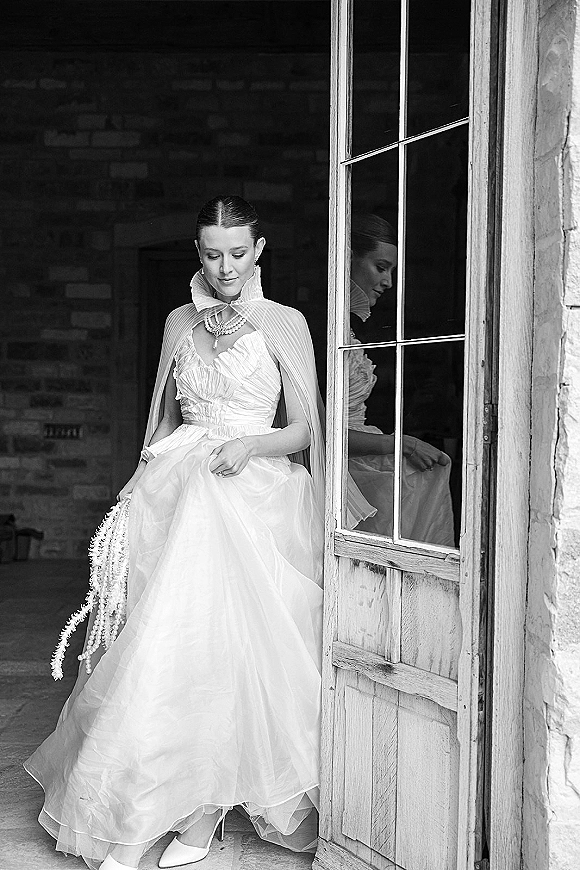 Bridal portrait in black and white of a bride looking down, wearing a high-neck ruffle gown with cape sleeves and pearls by a wooden door